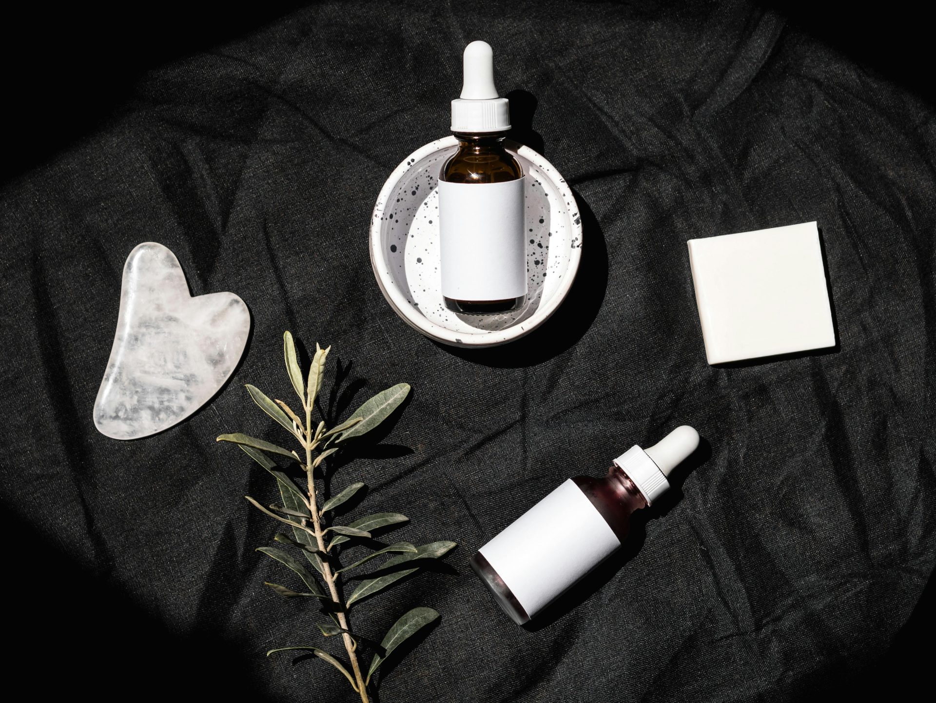 Stylish flat lay of skincare serum bottles with gua sha tool and leaves on a dark textile background.